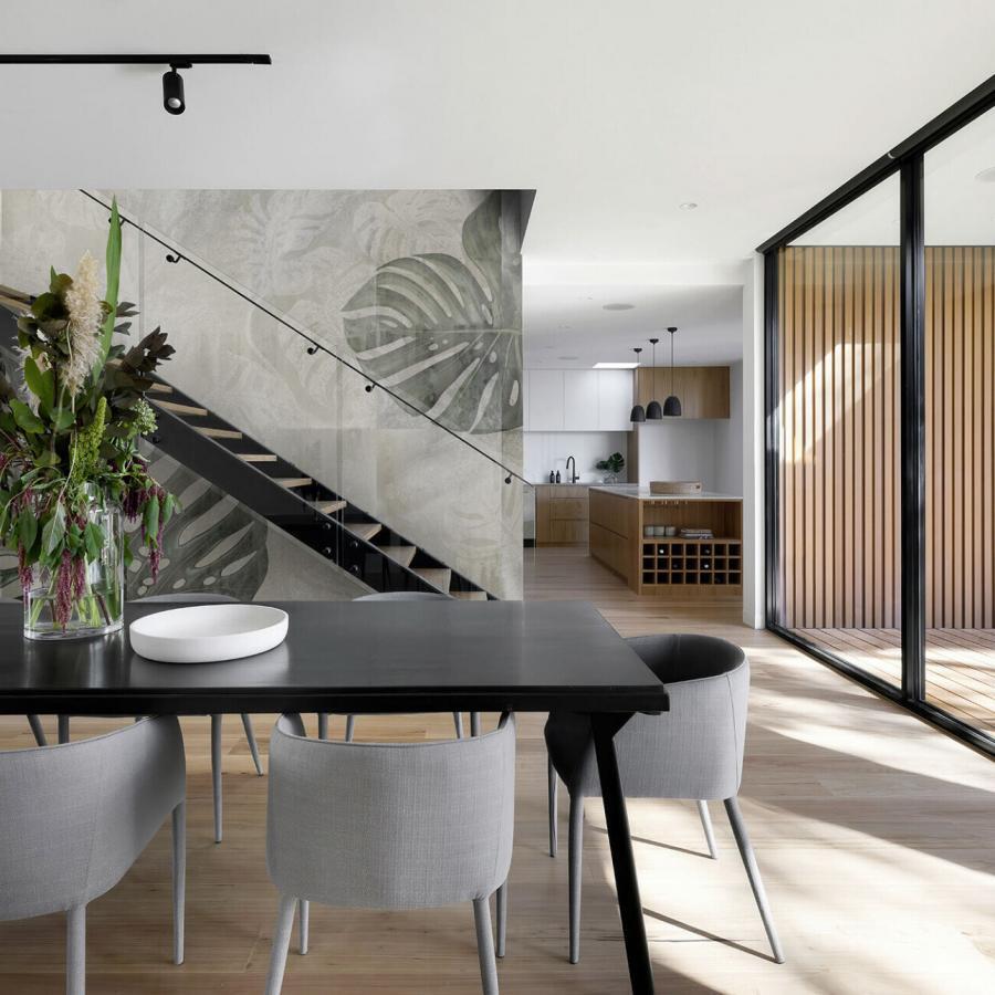 Monstera Tiles Sand Tapete in einem modernen Wohn- und Essbereich mit grauer Wand und großem Blättermuster, offener Küchenbereich und Glasfronten.