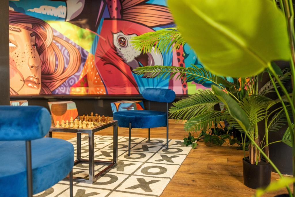 Blick in eine Lounge des Rioca Hotels in Frankfurt mit einer farbenfrohen Wand, blauen Velourssesseln, einem Schachtisch mit Schachfiguren und grünen Zimmerpflanzen
