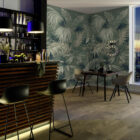 Tropische Blatttapete in Grün an einer Barwand mit Teakholz-Paneelen, beleuchtete Barhocker und eine Tischgruppe, entsprechend dem Heirloom-Wanddesign von k’moor