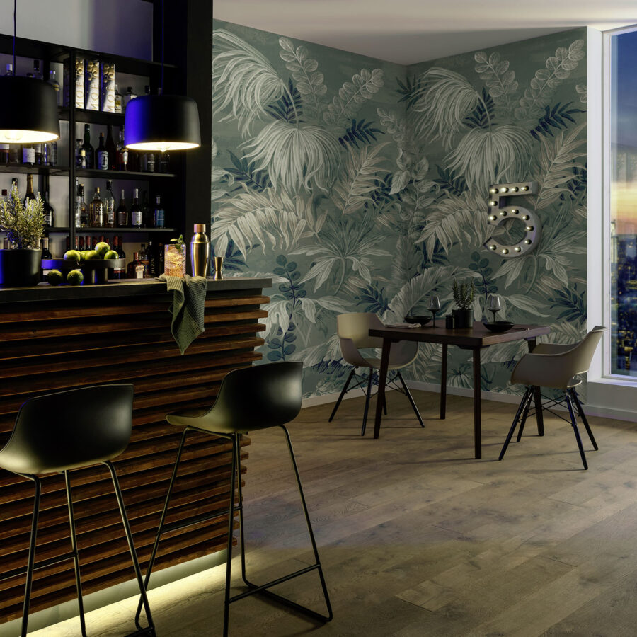 Tropische Blatttapete in Grün an einer Barwand mit Teakholz-Paneelen, beleuchtete Barhocker und eine Tischgruppe, entsprechend dem Heirloom-Wanddesign von k’moor