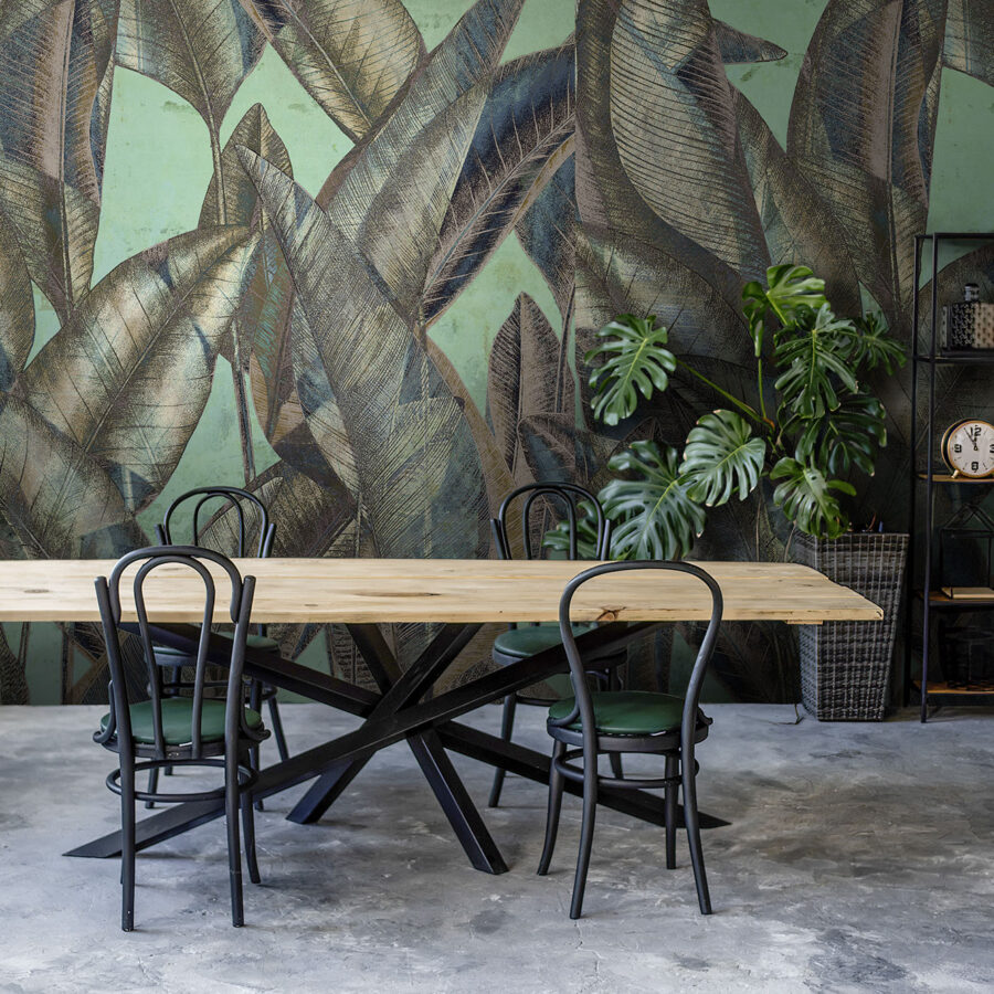 Wandtapete mit großem tropischen Blattmuster in Grüntönen vor einem Holztisch und schwarzen Metallstühlen, Teil des Believe-Designs von k’moor Wanddesign