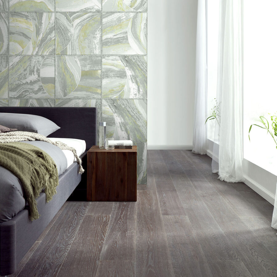 Milano Wanddesign in grau-grünen Fliesenmustern an der Schlafzimmerwand, links ein dunkelgraues Bett, daneben eine Holz-Nachtkommode, Boden aus dunklem Holz
