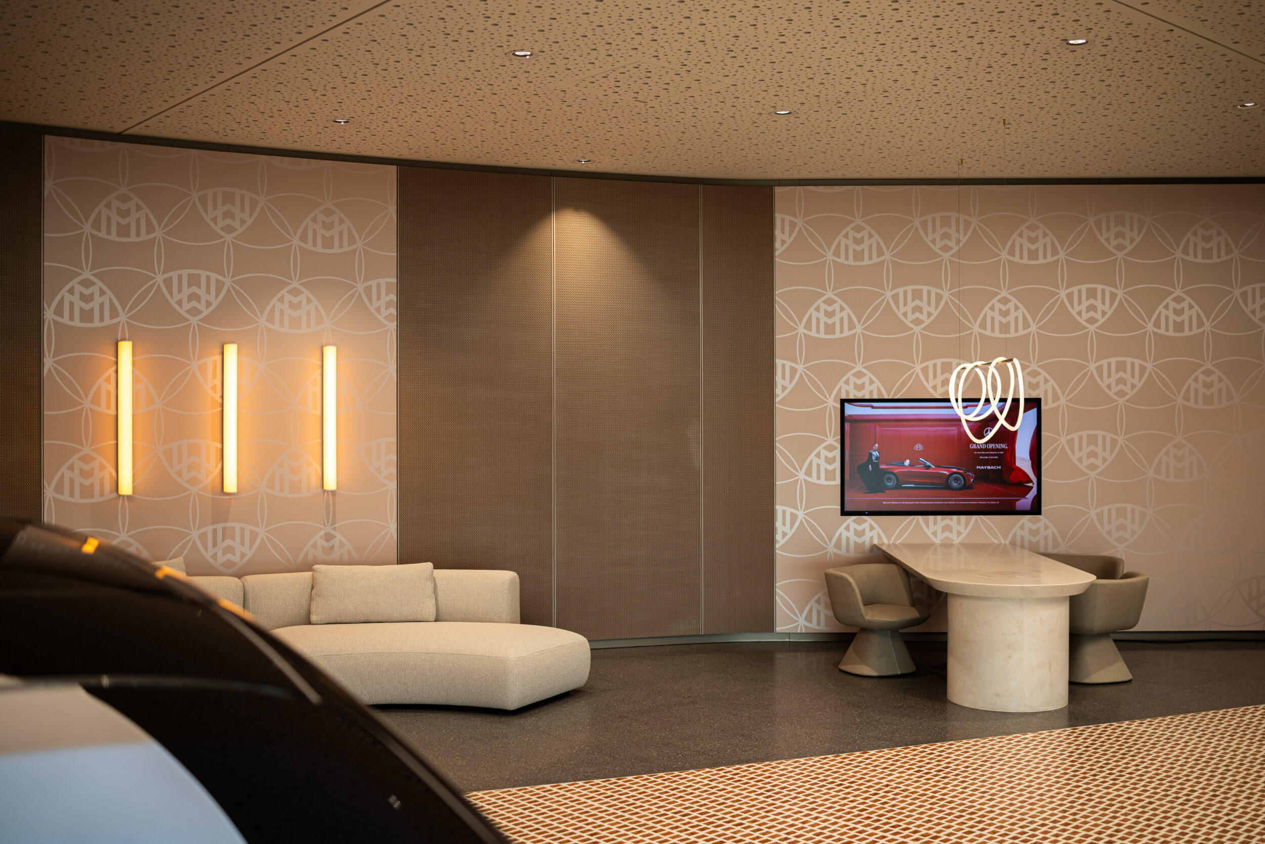 Tomislav Vukosav fotografierte die Tev Store Köln Lobby. Beige Musterwand, three vertical warmweiße Leuchten, komfortables Sofa, runder Tisch, Wand-TV