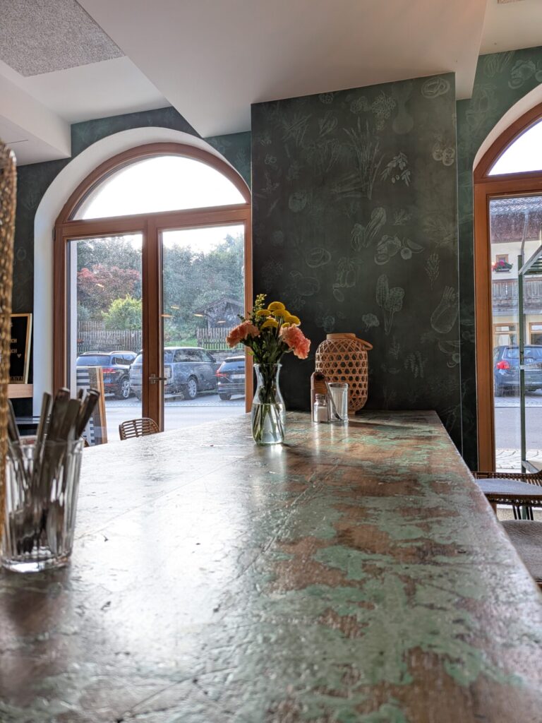 Café Stuerzer Innenraum mit dunkelgrüner Wandgestaltung, Holzrahmenfenster und stilvollem Barbereich; k’moor Wanddesign inspiriert von Veronika Lutz alias Vroni