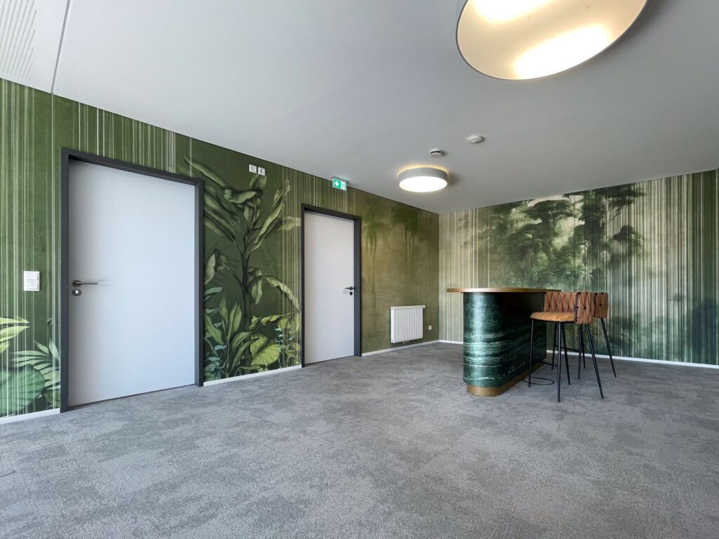 Green botanical wall designs and layered jungle motifs shape a modern office lounge with two white doors, grey carpet and a bar table.