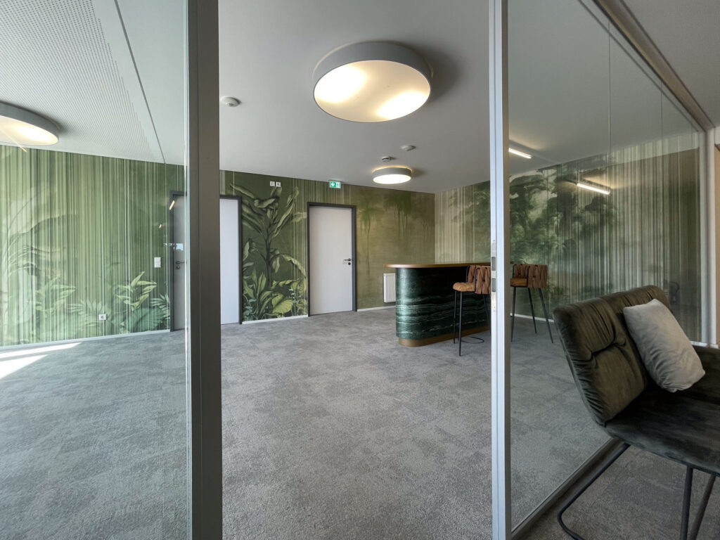 Ein grüner Büroraum mit tropischer Wandgestaltung, Glaswänden, Lounge-Sessel und Bartresen schafft eine ruhige, moderne Atmosphäre im Office.