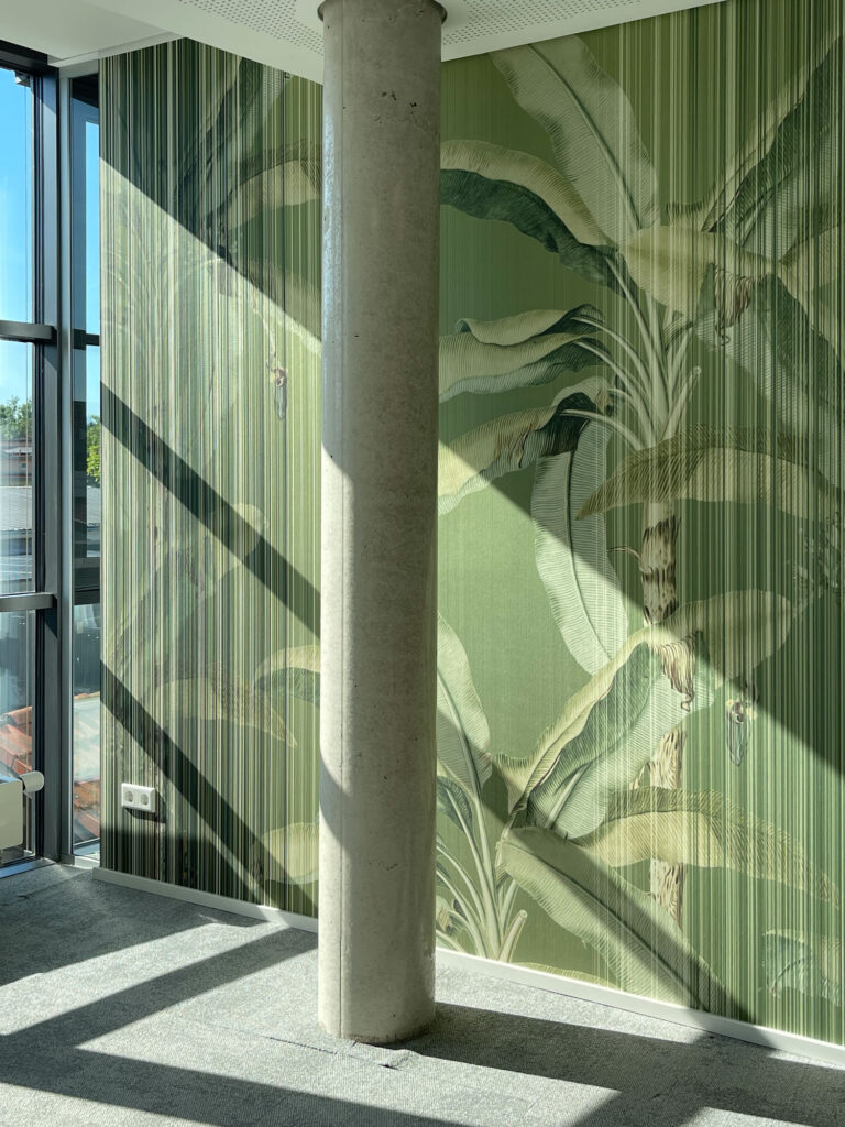 Im Büro trifft eine grüne botanische Wandgestaltung auf Betonstütze und große Fenster, während Sonnenlicht markante Schatten auf den Raum wirft.