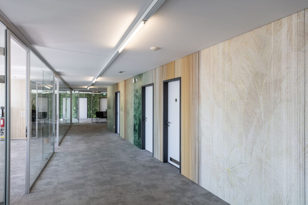 A long office corridor features k'moor wall design panels with botanical textures, vertical stripes, glass partitions and several white doors.
