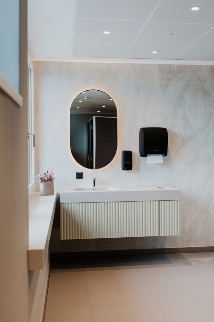 k-moor- Modernes Badezimmer mit ovalem Spiegel, schwebendem Waschtisch und Wanddesign Ein ovaler Spiegel über einem schwebenden Waschtisch trifft auf eine helle Betonoptik-Wand, schwarze Spender und dezente Beleuchtung.