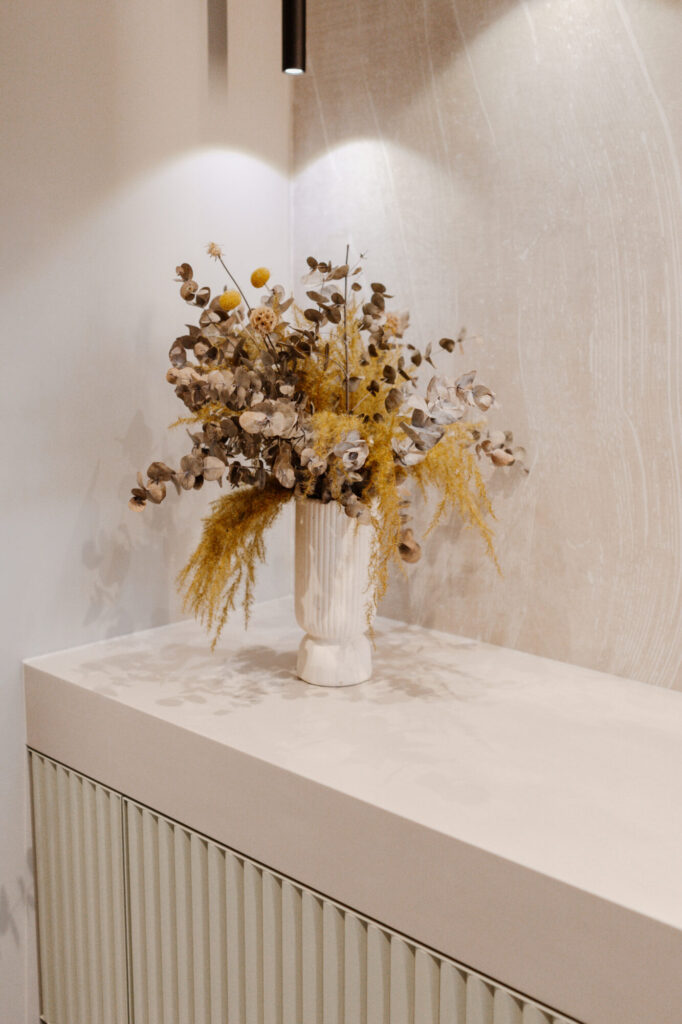 k-moor- Getrocknetes Blumenarrangement in geriffelter Vase auf hellem Sideboard, moderne Wandgestaltung Ein getrocknetes Blumenarrangement in einer geriffelten weißen Vase steht auf einem hellen Sideboard vor einer ruhigen, texturierten Wand.
