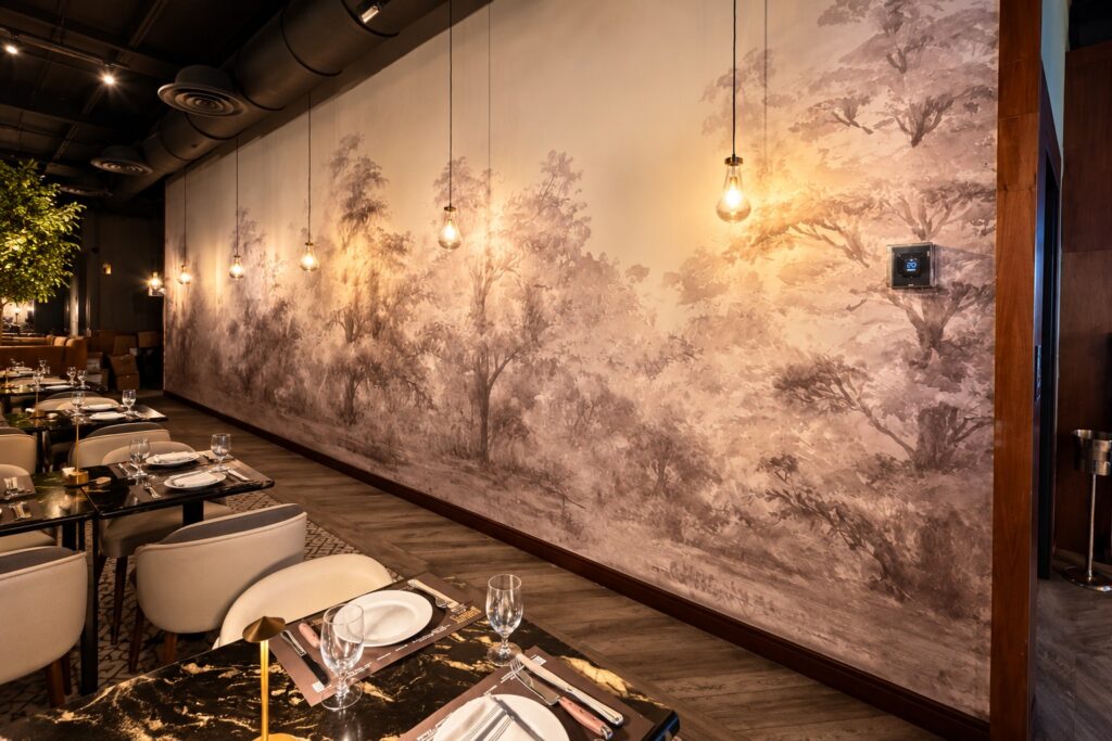 Gedeckte Tische und warm leuchtende Pendellampen vor dem sepiafarbenen Waldmotiv im Baires Grill in Miami.